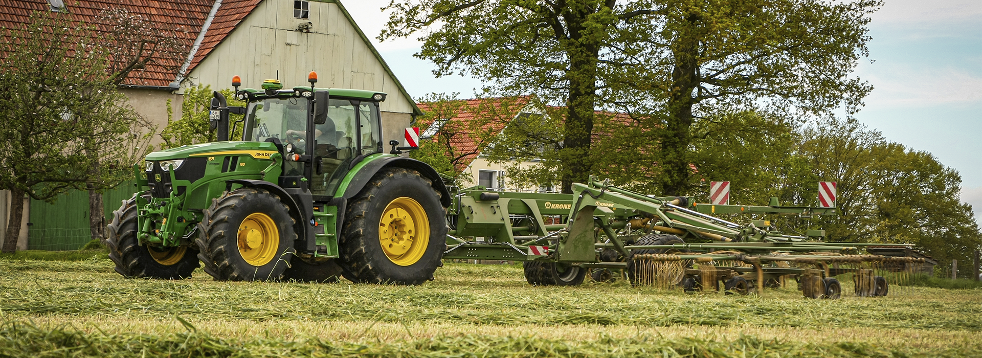 John Deere 6R 185 bei der Arbeit mit einem Krone Schwader vor einem alten Bauernhaus